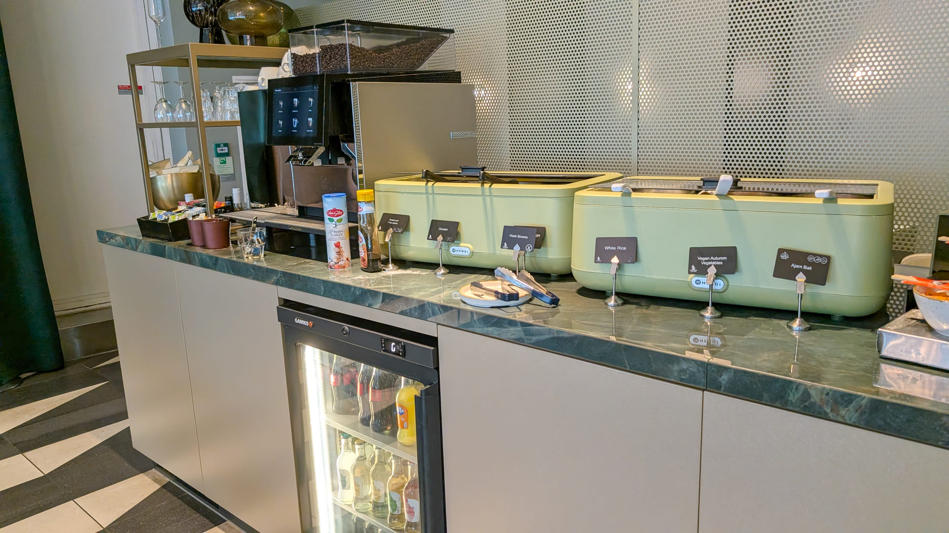 Ein moderner Buffetbereich in der Aspire Lounge mit einer professionellen Kaffeemaschine, hellgrünen Warmhaltebehältern für Speisen wie Reis und Gemüse sowie einem Untertischkühlschrank mit Kaltgetränken.
