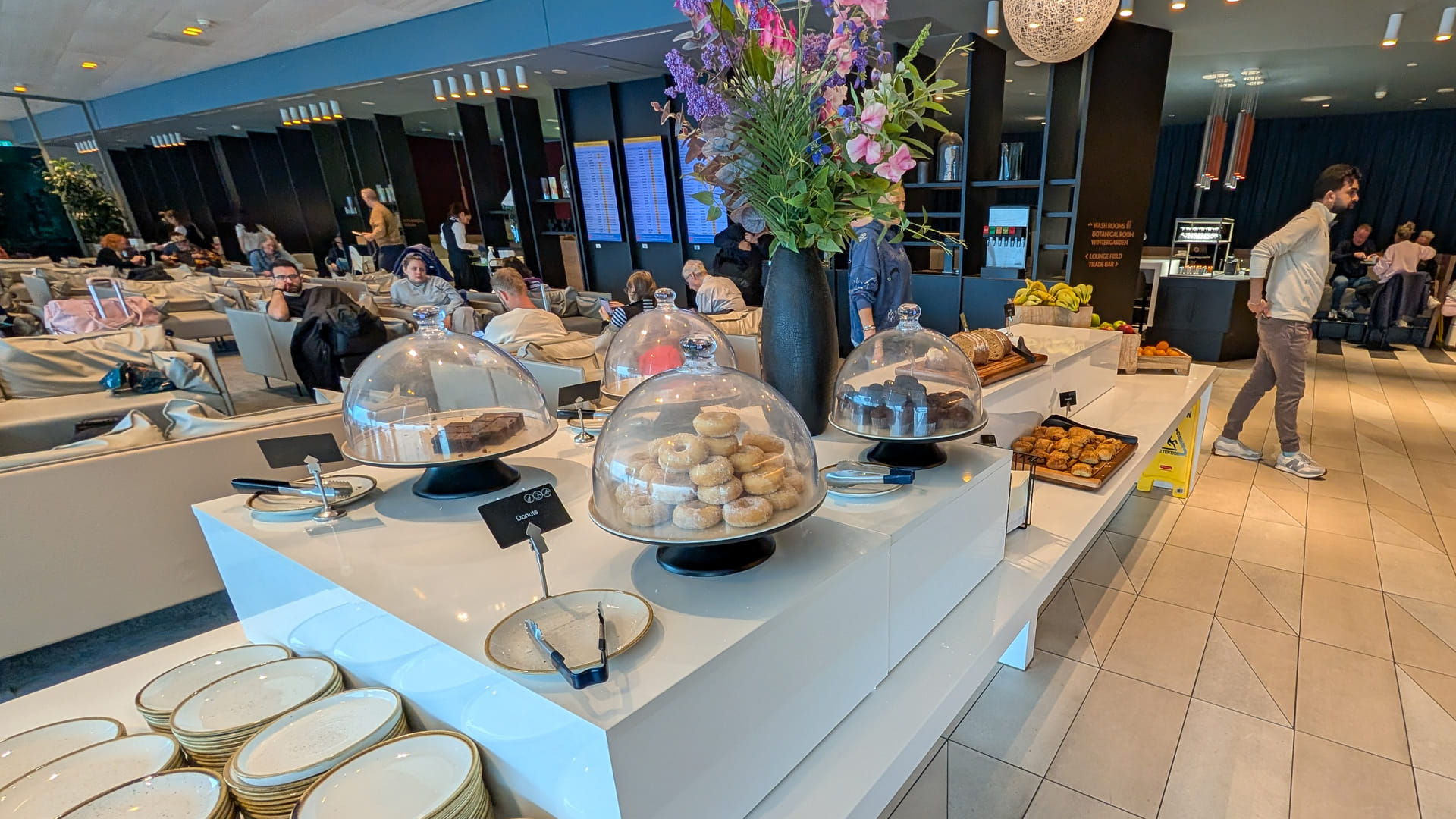 Buffetstation in einer Flughafenlounge mit Glasglocken über Donuts, Gebäck und Aufschnitt, im Hintergrund ein Sitzbereich mit Passagieren.