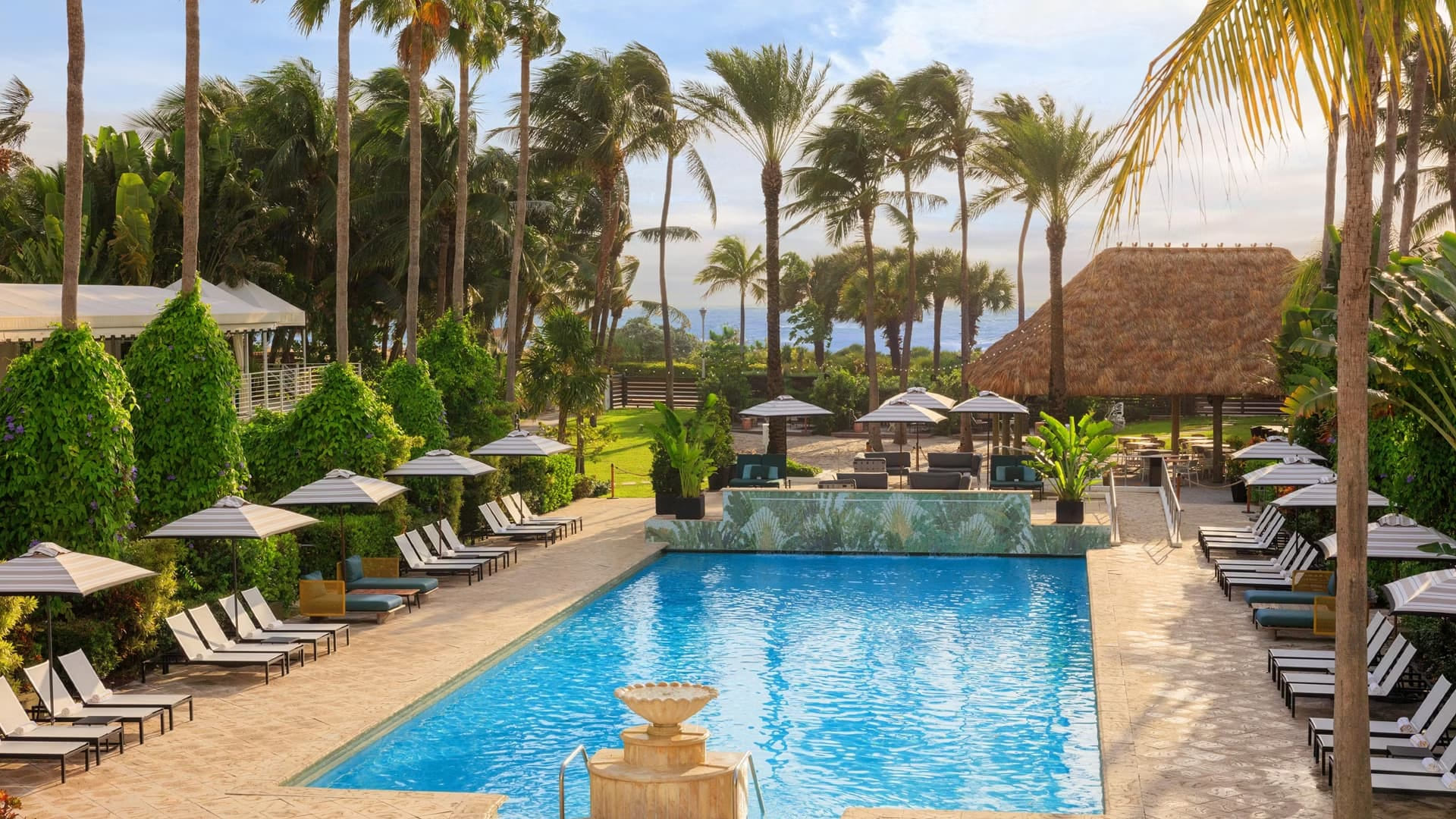 Blick auf den Außenpool des Kimpton Surfcomber Hotels in Miami Beach. Im Vordergrund glitzert blaues Poolwasser vor einer mit Palmenmotiven gefliesten Wand. Dahinter schließt sich ein Loungebereich mit modernen Gartenmöbeln, gestreiften Sonnenschirmen und Palmen an, der direkt in einen sandigen Bereich übergeht. Im Hintergrund sind das Meer und ein sonniger Himmel unter Palmen zu erkennen.