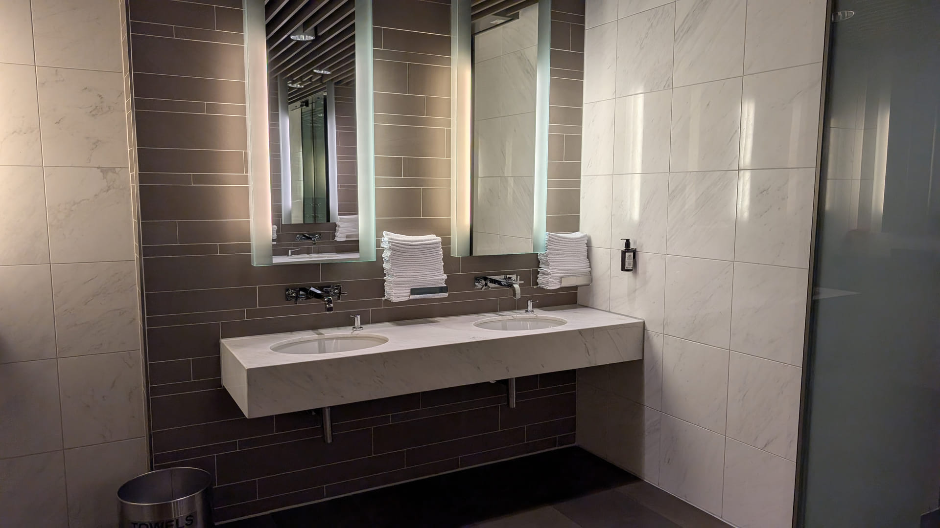 Ein modernes, luxuriöses Badezimmer im Lufthansa First Class Terminal. Ein Doppelwaschtisch aus weißem Marmor ist vor einer dunkelgrauen Fliesenwand installiert. Über den Waschbecken hängen zwei große, vertikale Spiegel mit integrierter LED-Hintergrundbeleuchtung. Zwischen und neben den Spiegeln liegen ordentlich gestapelte, weiße Stoffhandtücher bereit. Die restlichen Wände sind mit großformatigen, hellen Marmorfliesen gestaltet, was dem Raum eine helle und sterile Eleganz verleiht.
