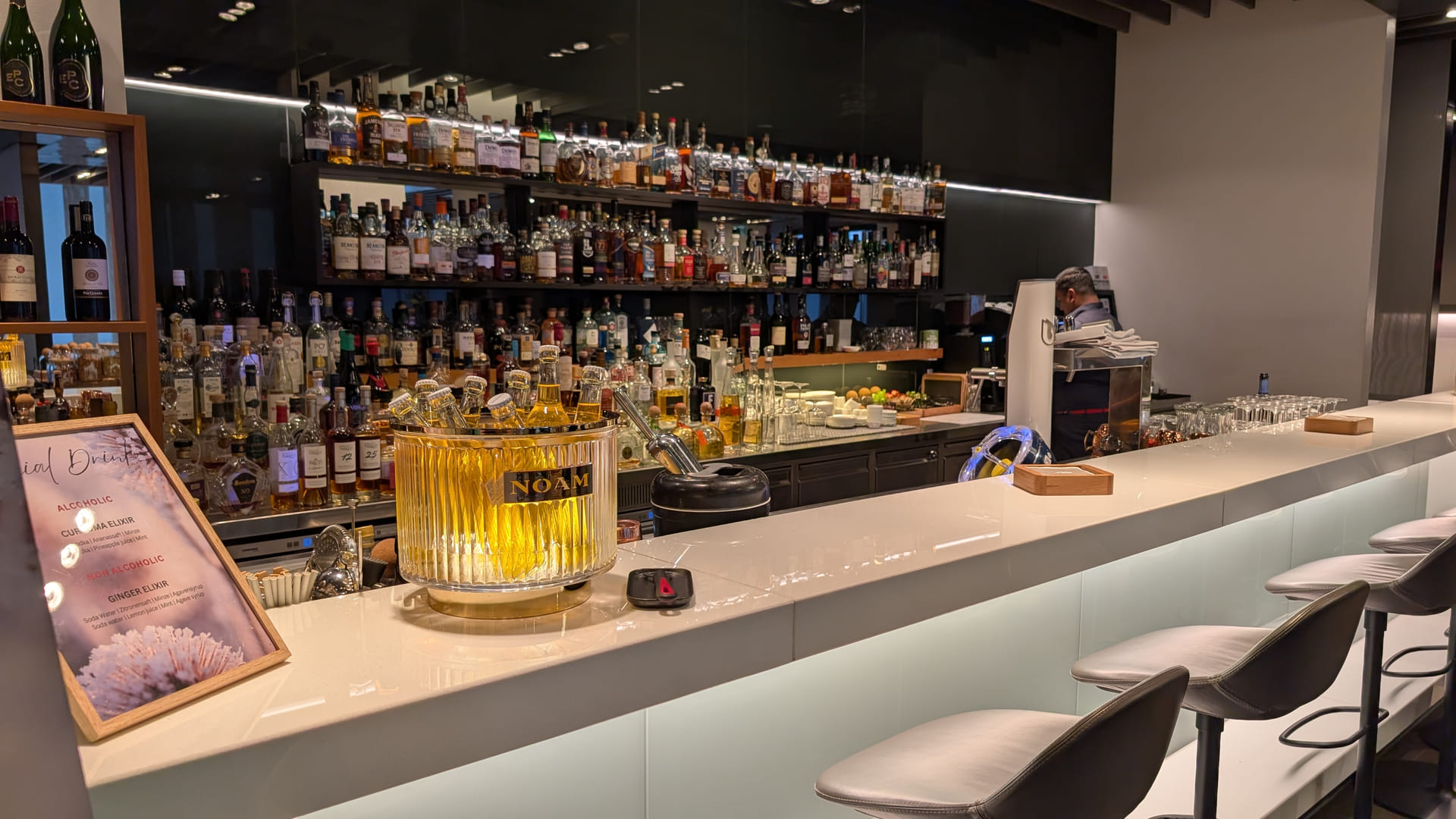 Eine weitläufige, modern beleuchtete Bar im Lufthansa First Class Terminal. Die Regale im Hintergrund sind mit einer beeindruckenden Auswahl an hochwertigen Spirituosen und Weinen gefüllt. Auf dem weißen Tresen steht ein beleuchteter Glas-Eiskübel mit „Noam“-Bierflaschen. Im Vordergrund ist eine Getränkekarte mit „Special Drinks“ zu sehen, während rechts elegante Barhocker aus grauem Leder zur Entspannung einladen.