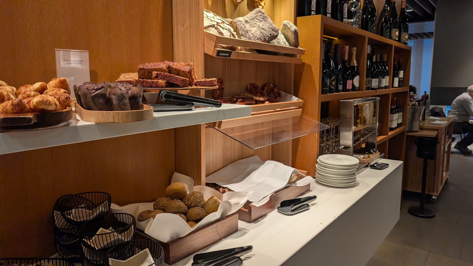 Ein hölzernes Buffetregal mit einer Auswahl an frischem Gebäck. In den oberen Fächern liegen rustikale Brotlaibe und Brezeln, darunter befinden sich Körbe mit verschiedenen Brötchen sowie Tabletts mit Croissants, Muffins und Kuchenstücken. Im Hintergrund sind Weinflaschen in einem Regal aufgereiht.