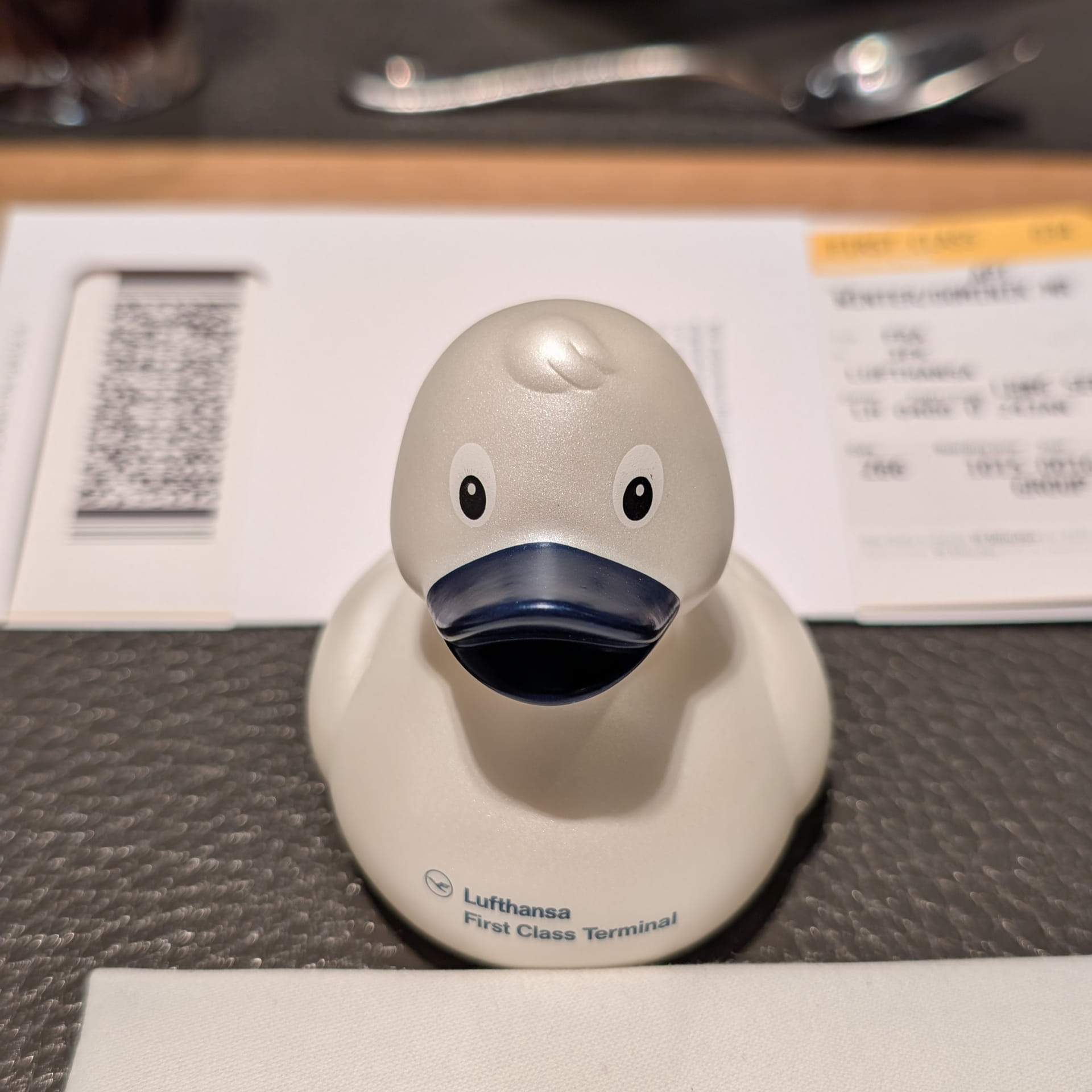 Nahaufnahme einer cremeweißen Quietscheente mit dunkelblauem Schnabel, einem Sammlerstück aus dem Lufthansa First Class Terminal. Auf der Brust der Ente ist dezent das Lufthansa-Logo und der Schriftzug „Lufthansa First Class Terminal“ aufgedruckt. Die Ente steht auf einem grauen Tisch, im unscharfen Hintergrund ist eine Bordkarte erkennbar.