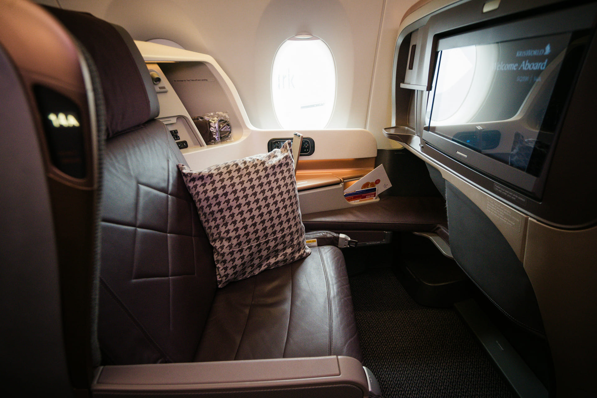 Singapore Airlines Business Class Sitz im Airbus A350 mit braunem Ledersitz, Hahnentritt-Kissen und großem HD-Bildschirm mit KrisWorld Welcome-Aboard-Anzeige am Fenster.
